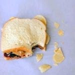 A peanut butter and jelly sandwich with potato chips with one bite gone and some chips scattered around it.
