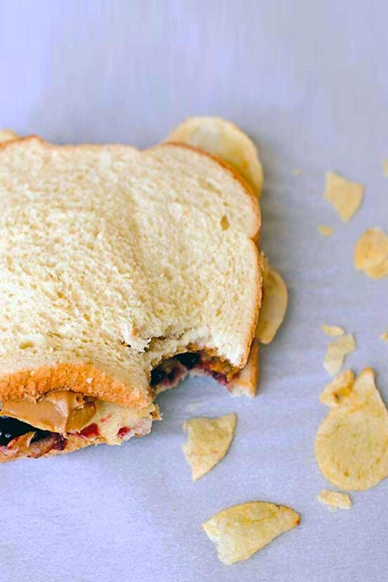 A peanut butter and jelly sandwich with potato chips with one bite gone and some chips scattered around it.
