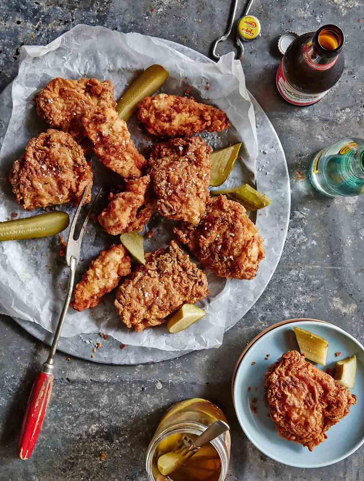 A platter of pickle-brined fried chicken with pieces of dill pickle, and a bottle of beer.