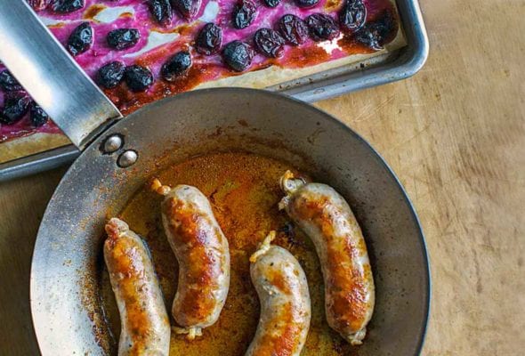 Skillet with four seared pork sausages with roasted grapes on a baking sheet nearby.