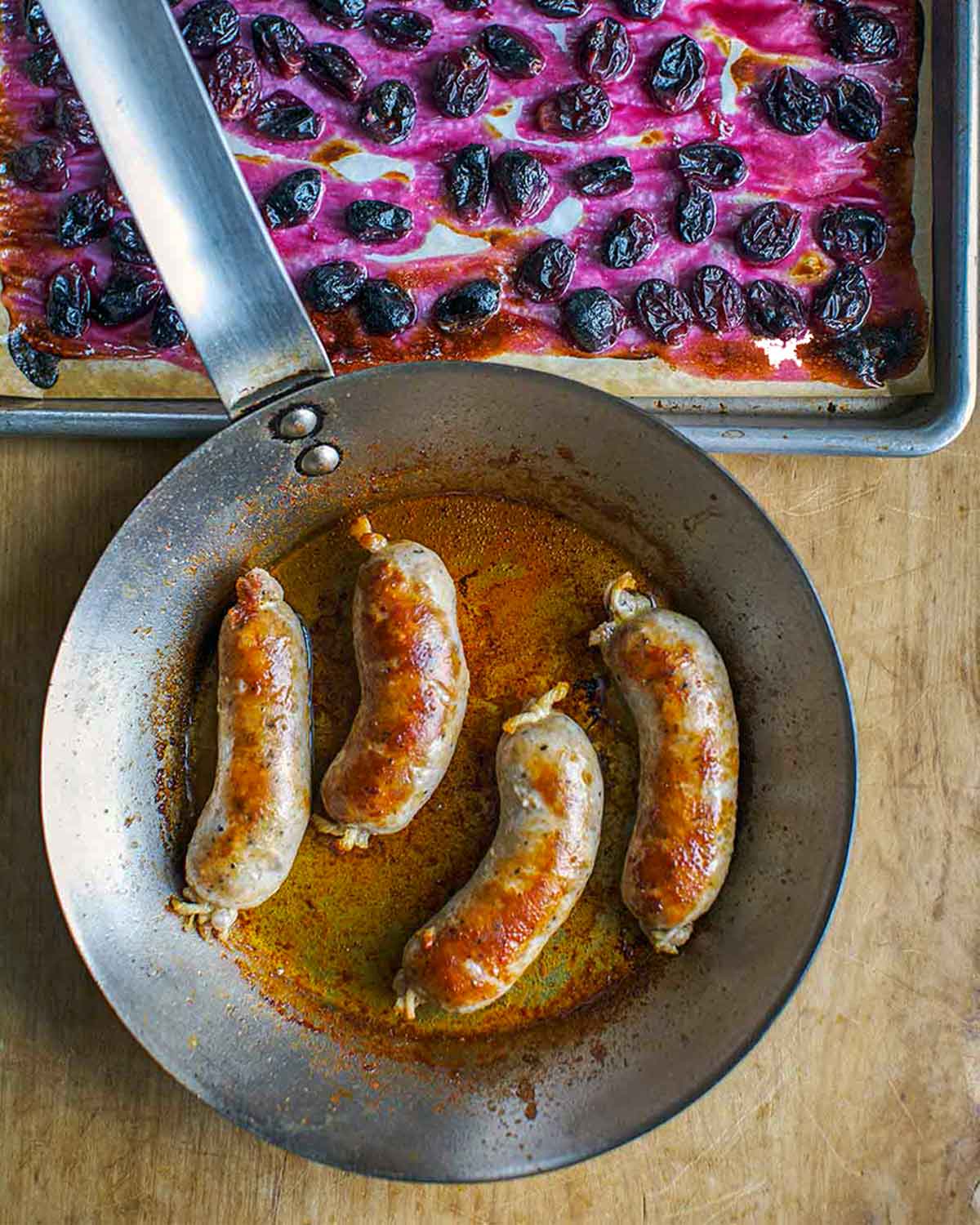 Skillet with four seared pork sausages with roasted grapes on a baking sheet nearby.