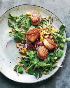 A plate of seared scallops with grilled sweet corn salad, sliced red onion, and watercress.