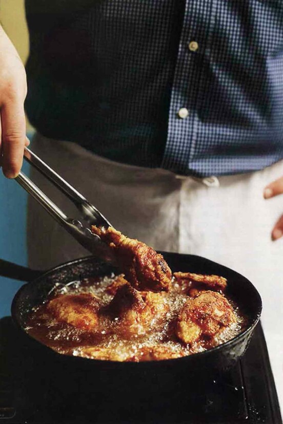 A man holding tongs in front of a skillet making southern pan-fried chicken, which is bubbling in the pan