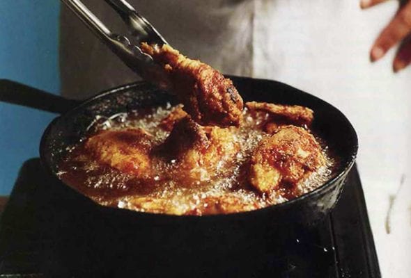 A man holding tongs in front of a skillet making southern pan-fried chicken, which is bubbling in the pan.
