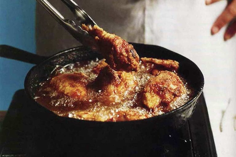 A man holding tongs in front of a skillet making southern pan-fried chicken, which is bubbling in the pan.
