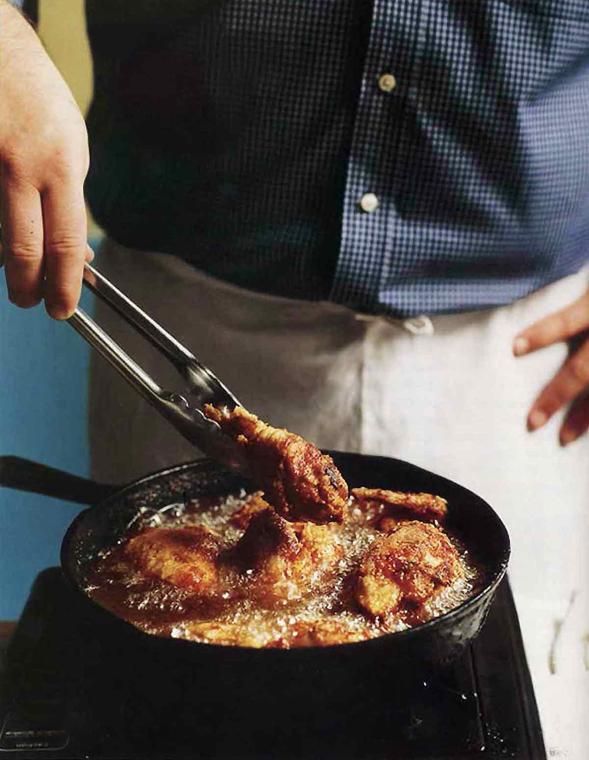 A man holding tongs in front of a skillet making southern pan-fried chicken, which is bubbling in the pan