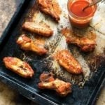 A baking sheet with 6 spicy baked chicken wings glazed with hot sauce, a jar of sauce nearby