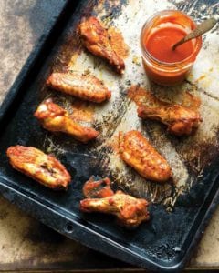 A baking sheet with 6 spicy baked chicken wings glazed with hot sauce, a jar of sauce nearby