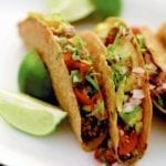 Three sweet and spicy tacos on a white plate with wedges of lime alongside.