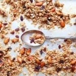 Almond and coconut granola scattered on a baking sheet with a spoon resting in the center.