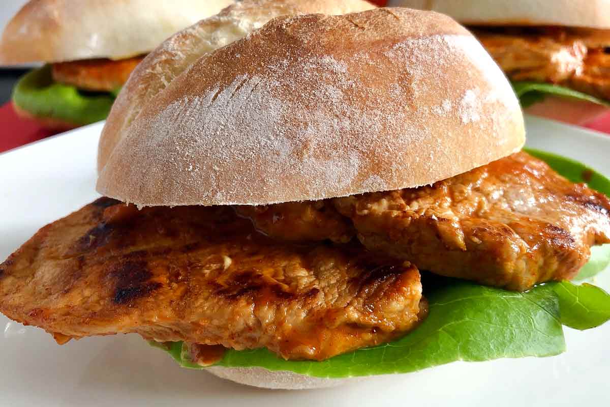 An assembled bifana with lettuce on a papo secos roll on a white plate.