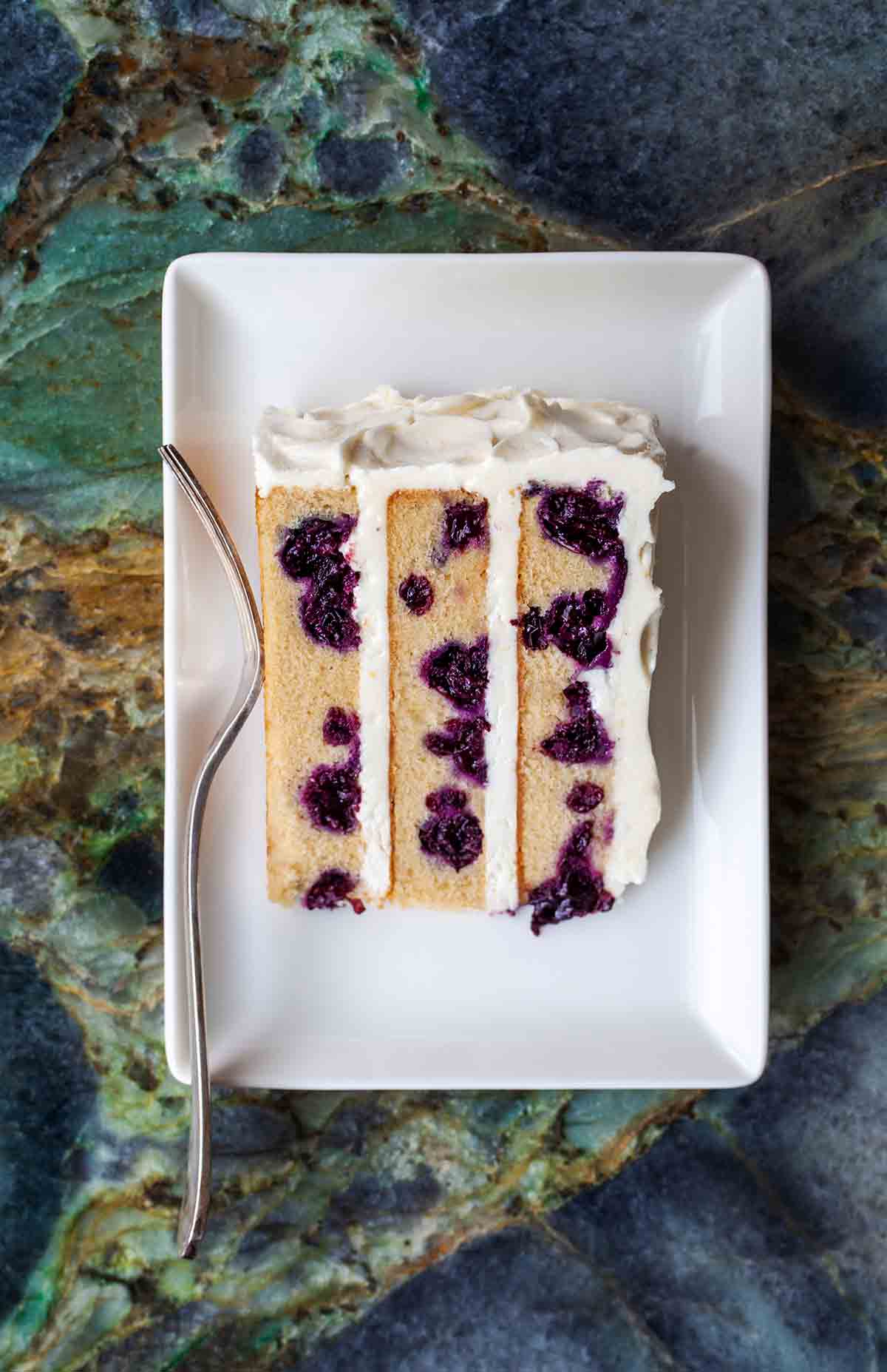 A white plate with a slice of blueberry-lemon layer cake.