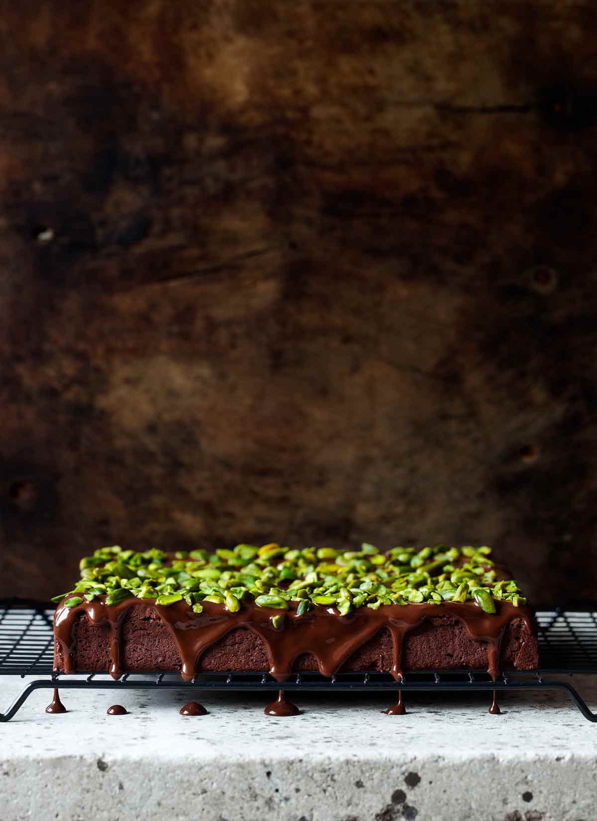 A slab of chocolate brownies with chocolate drizzle and chopped pistachios made by Cenk Sönmezsoy on a wire rack.