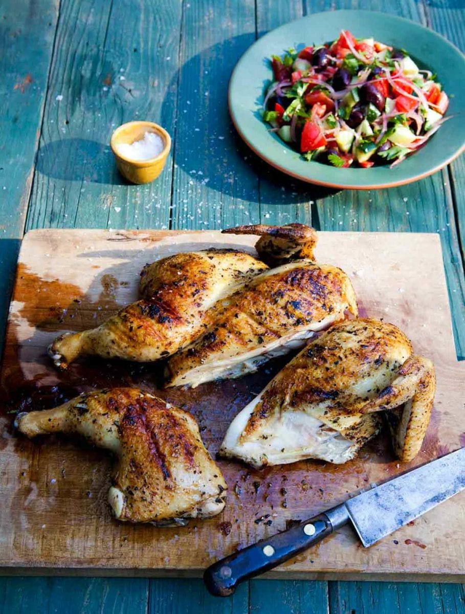 A wooden cutting board with a carved grilled Greek chicken and a knife resting beside it.