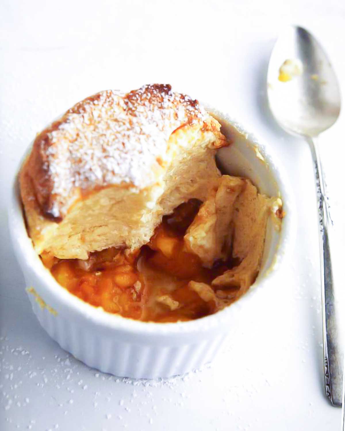 White dish of a sugar-dusted individual peach souffle; in the bottom are peach slices