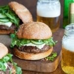 Three lamb burgers with arugula and feta on a wooden surface with two jars of beer beside them.