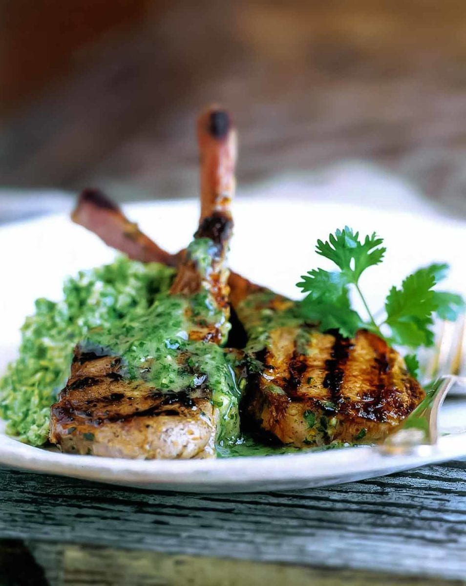 Two grilled lamb chops with cilantro-mint sauce on a white plate with a fork and a sprig of cilantro to garnish.