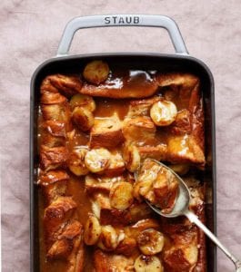 A Staub casserole dish filled with overnight French toast with caramel sauce and bananas and spoon resting in the dish.
