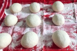 Nine balls of dough rising on a towel-lined baking sheet for papo-secos.