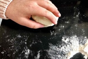A person rolling a ball of dough for papo-secos on a flour-dusted black surface.