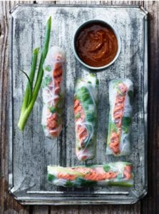Four salmon summer rolls on a metal sheet with a piece of scallion and a bowl of dipping sauce.