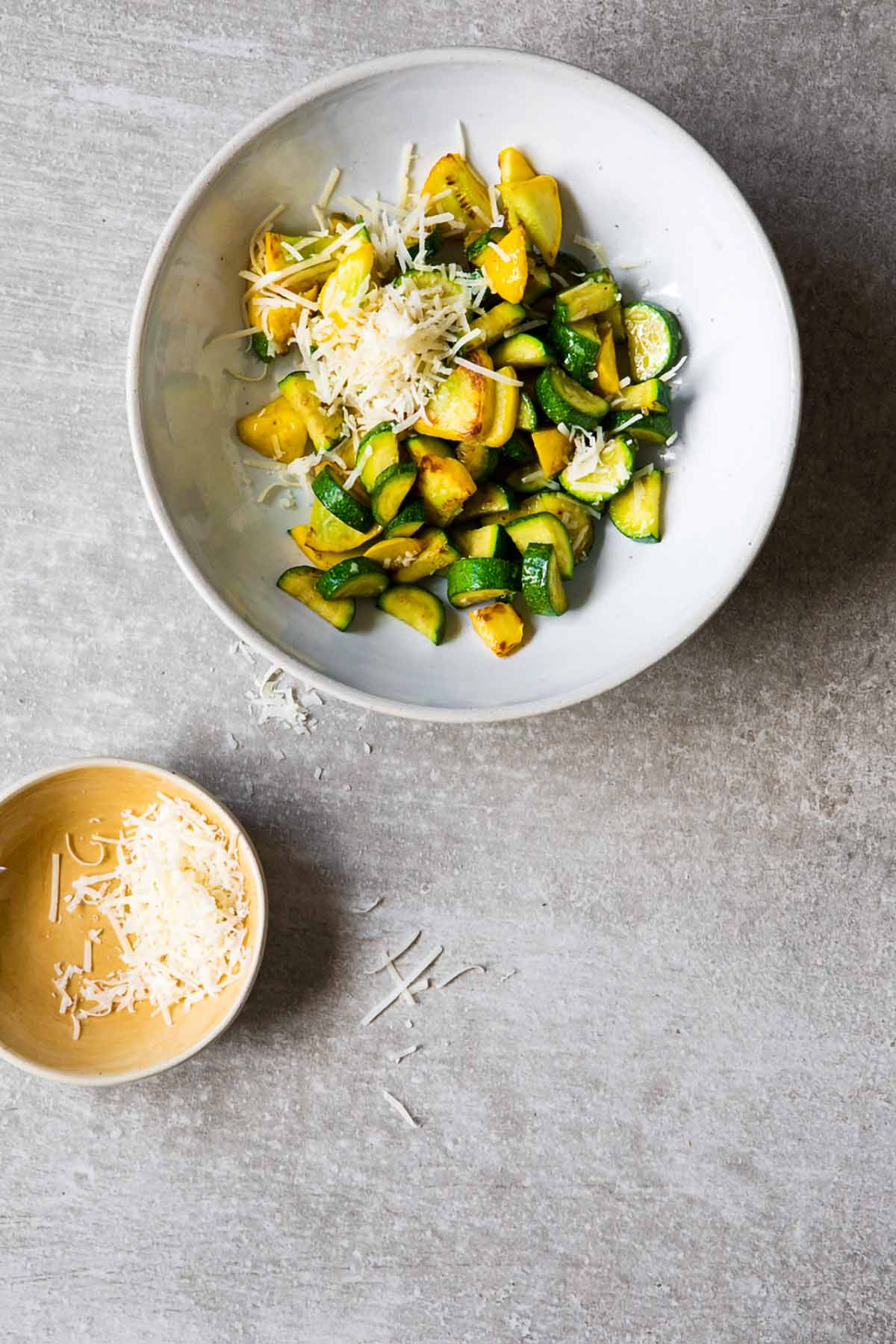 A white bowl filled with sautéed summer squash with a sprinkle of Parmesan on top and a small bowl of Parmesan on the side.