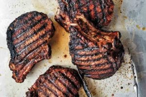 Four spice-rubbed grilled pork chops on a baking sheet with a pair of metal tongs