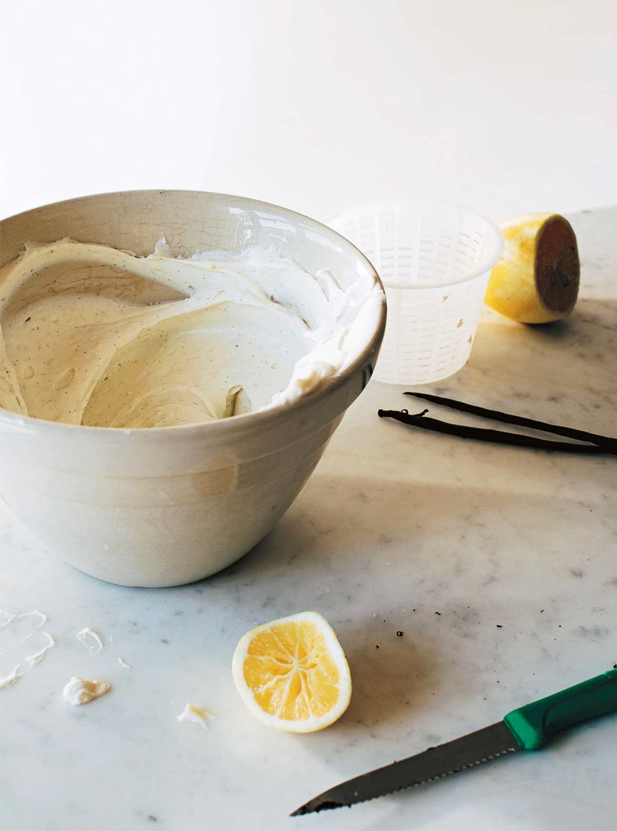 A bowl of sweet whipped ricotta cream, a halved lemon, two vanilla beans, a knife, and a small glass on a marble surface.