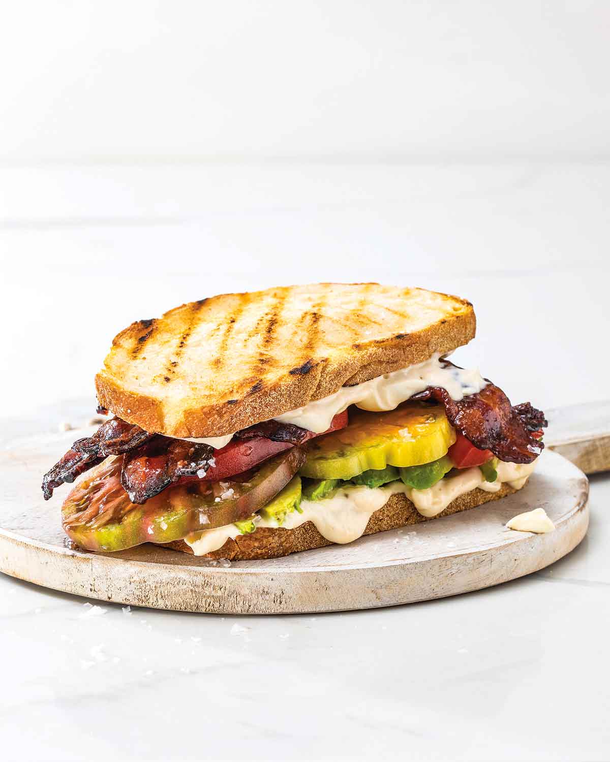 A bacon avocado tomato sandwich on a round wooden board.