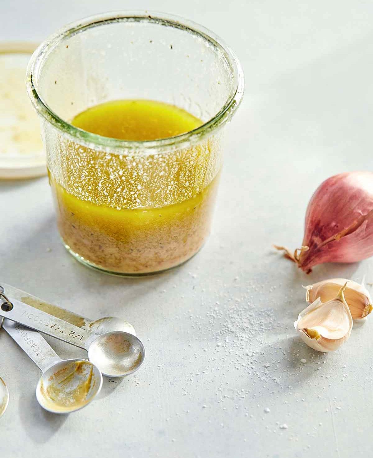 A glass jar half-filled with basic vinaigrette with measuring spoons, a shallot, and two garlic cloves on the side.