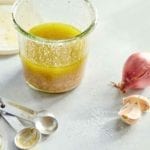 A glass jar half-filled with basic vinaigrette with measuring spoons, a shallot, and two garlic cloves on the side.