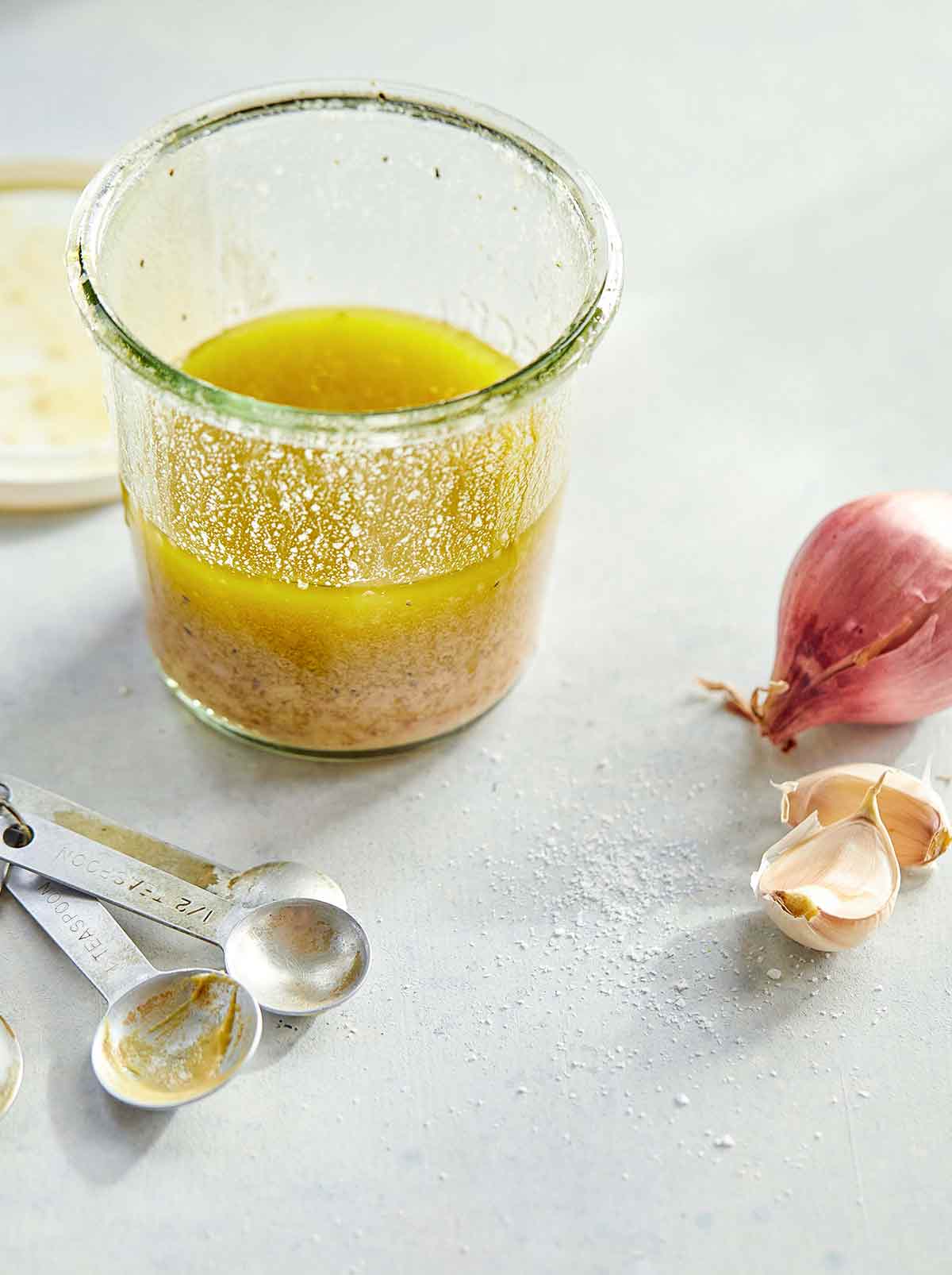 A glass jar half-filled with basic vinaigrette with measuring spoons, a shallot, and two garlic cloves on the side.