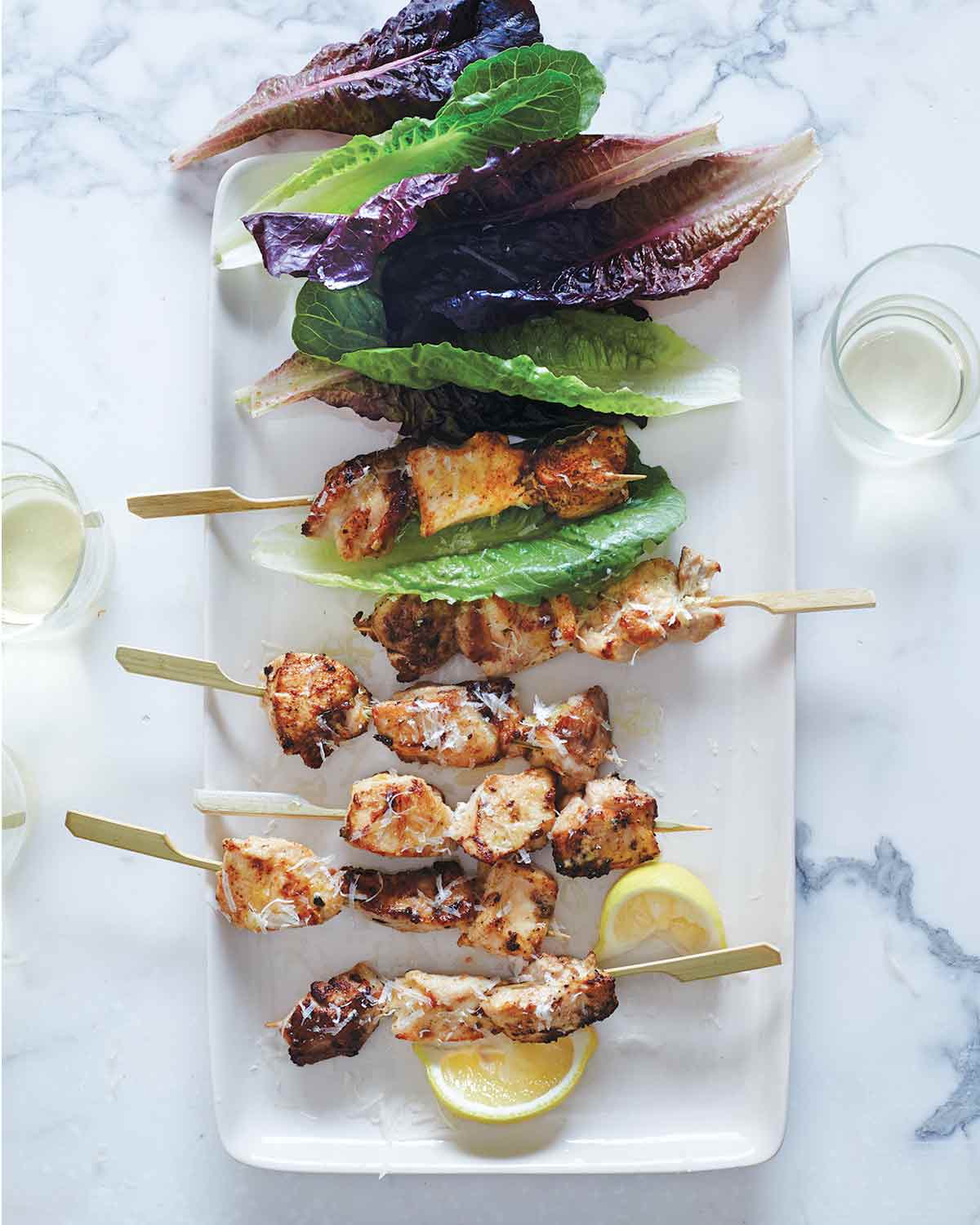 Six chicken caesar salad skewers on a white plate with leaves of red and green romaine and two lemon wedges.