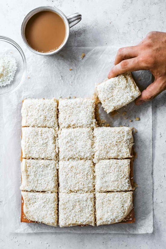 A frosted coconut sheet cake cut into 12 squares on a piece of parchment with a person grabbing a piece.