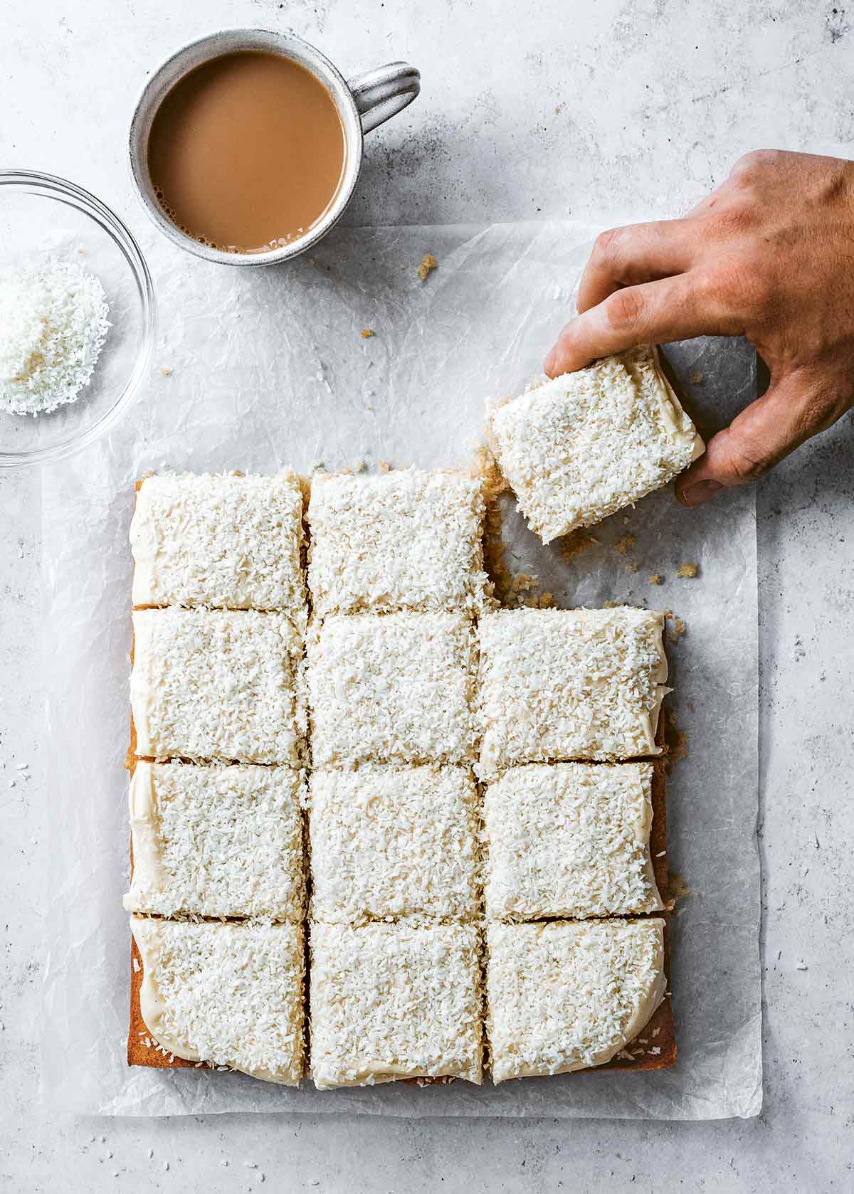 A frosted coconut sheet cake cut into 12 squares on a piece of parchment with a person grabbing a piece.