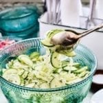 A serving of cucumber and onion salad being lifted from a serving bowl with silver salad tongs.