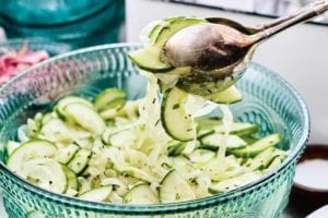 A serving of cucumber and onion salad being lifted from a serving bowl with silver salad tongs.