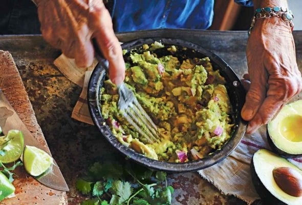 A person mashing avocado into a bowl of easy guacamole on a table with salt, avocado, cilantro, lime, and chiles.