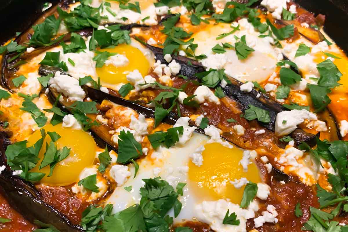 A close view of eggplant shakshuka made with poached eggs in a tomato and eggplant sauce, with cilantro and feta scattered over the top.