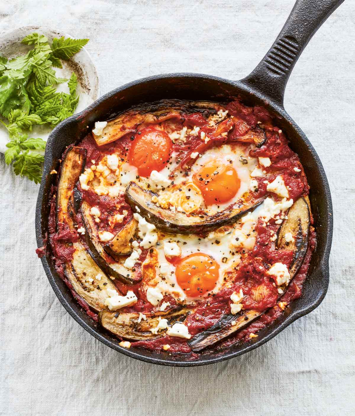 A cast-iron skillet filled with eggplant shakshuka made with poached eggs in a tomato and eggplant sauce, with feta scattered over the top.