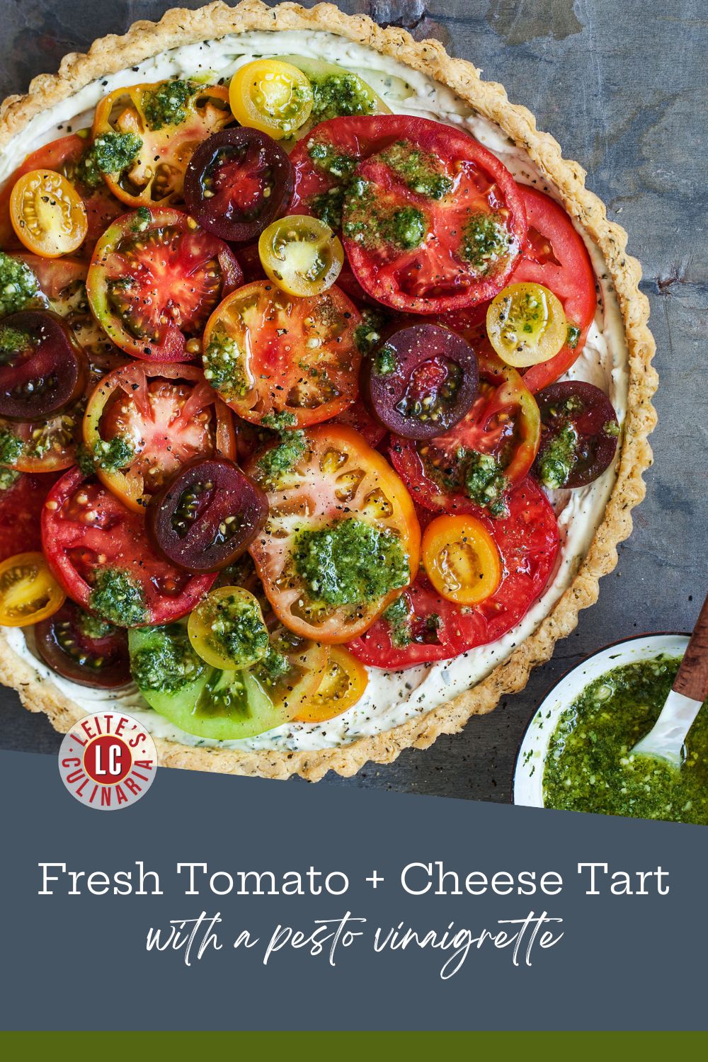 A colorful, fresh heirloom tomato tart on a gray slate background, with a small white bowl of pesto vinaigrette on the side.