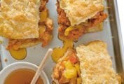 Three fried chicken and biscuits sandwiches on a baking sheet with a bowl of honey beside and drizzled around the sandwiches.