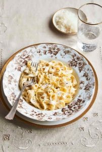 A patterned bowl filled with lemony fettuccine alfredo on a white and gold plate with a glass of water and dish of Parmesan on the side.