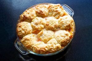 A round glass baking dish filled with peach nectarine cobbler and a spoon on the side.