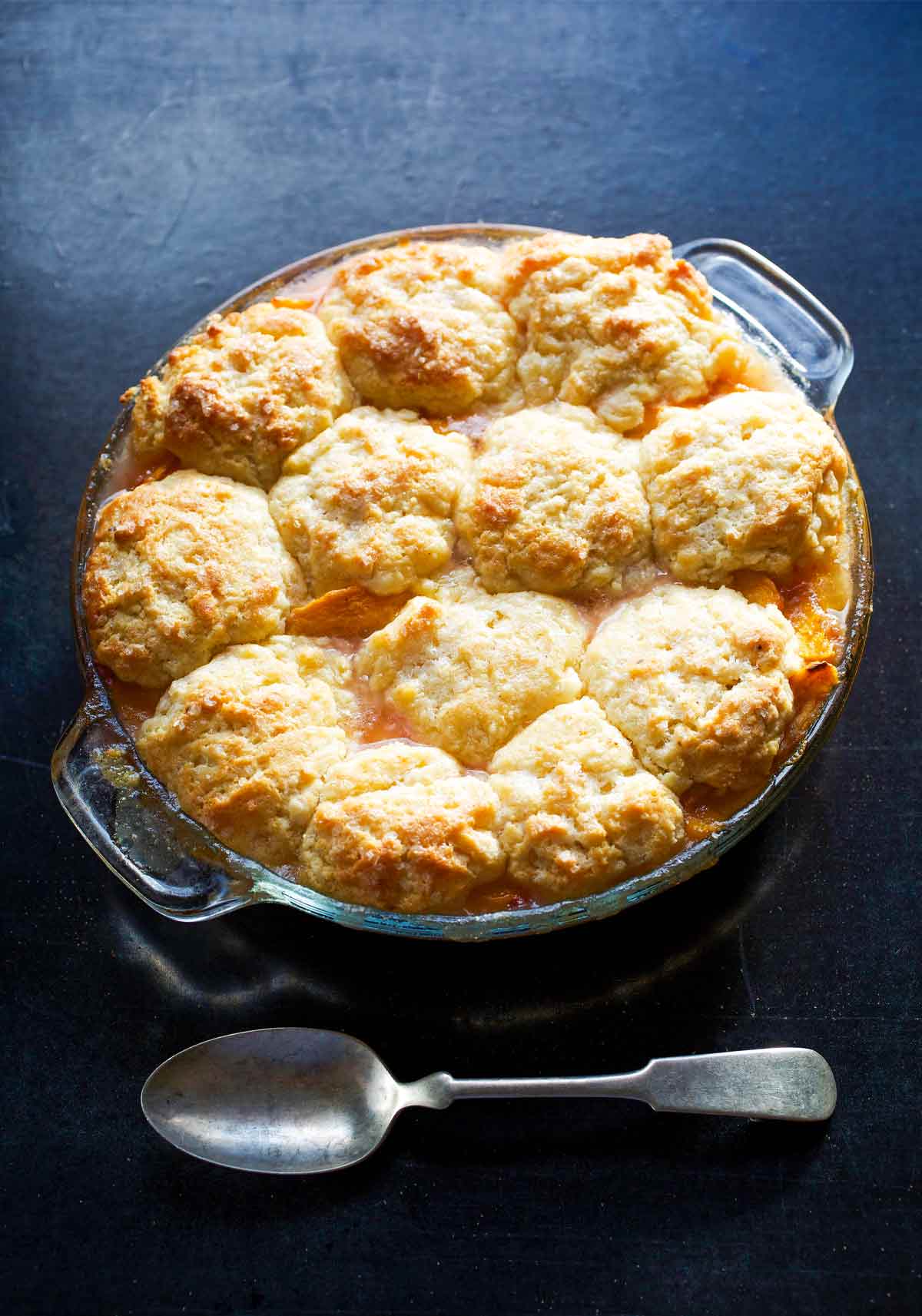 A round glass baking dish filled with peach nectarine cobbler and a spoon on the side.