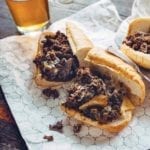 Two Philly cheesesteak sandwiches cut in half on a foil wrapper with a glass of beer in the background.