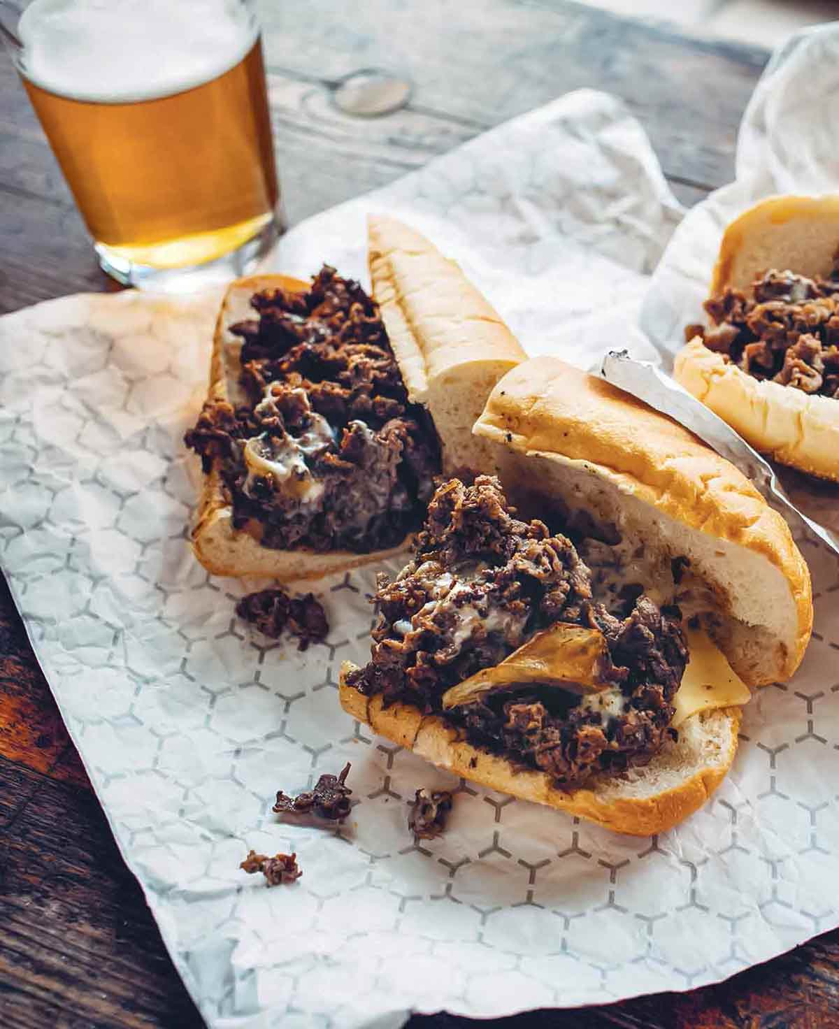 Two Philly cheesesteak sandwiches cut in half on a foil wrapper with a glass of beer in the background.