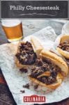 Two Philly cheesesteak sandwiches cut in half on a foil wrapper with a glass of beer in the background.