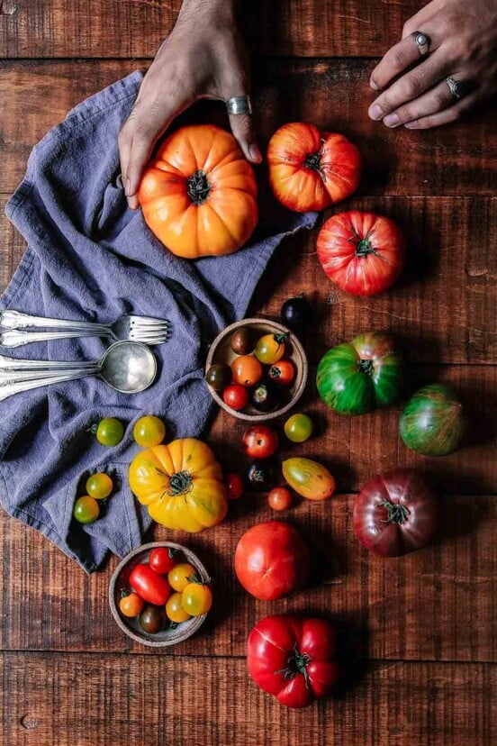 A wooden table filled with assorted tomatoes, a cloth napkin, spoons, and forks, and a person gently squeezing a tomato to check if it is perfectly ripe.