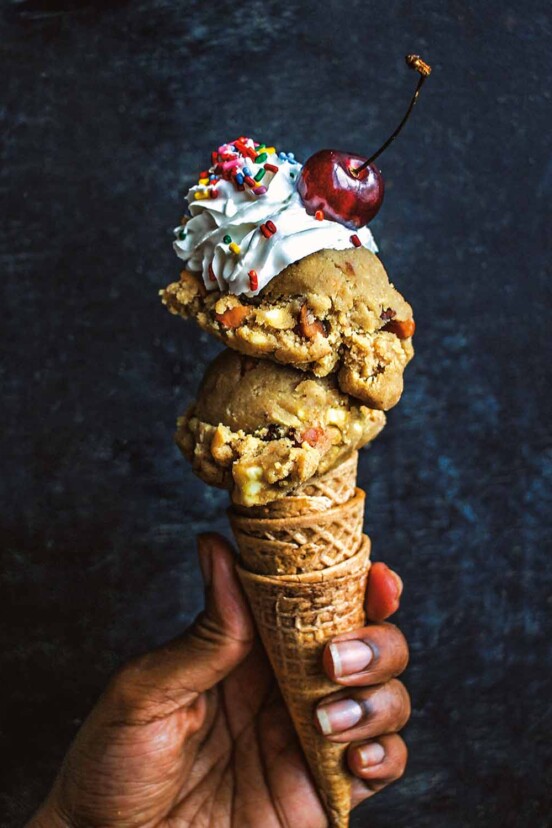 A person holding three nested ice cream cones topped with two scoops of vegan gluten-free chocolate chip cookie dough, whipped cream, sprinkles, and a cherry.